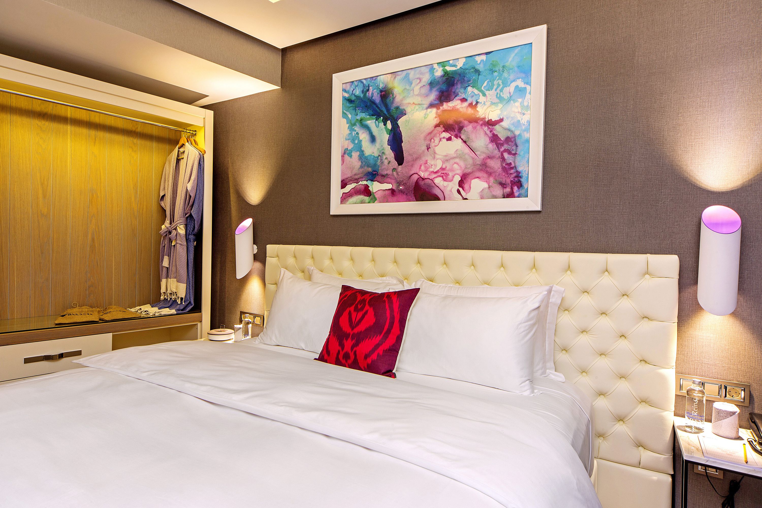 Ein Hotelzimmer mit einem großen Bett, einem Kleiderhaken mit einem Bademantel, einem Gemälde an der Wand und einem Nachttisch mit Lampe und Telefon.