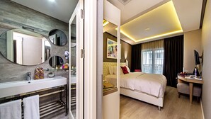 Habitació d'hotel moderna amb un llit gran, una taula de nit, un escriptori i un bany amb un lavabo i un mirall.