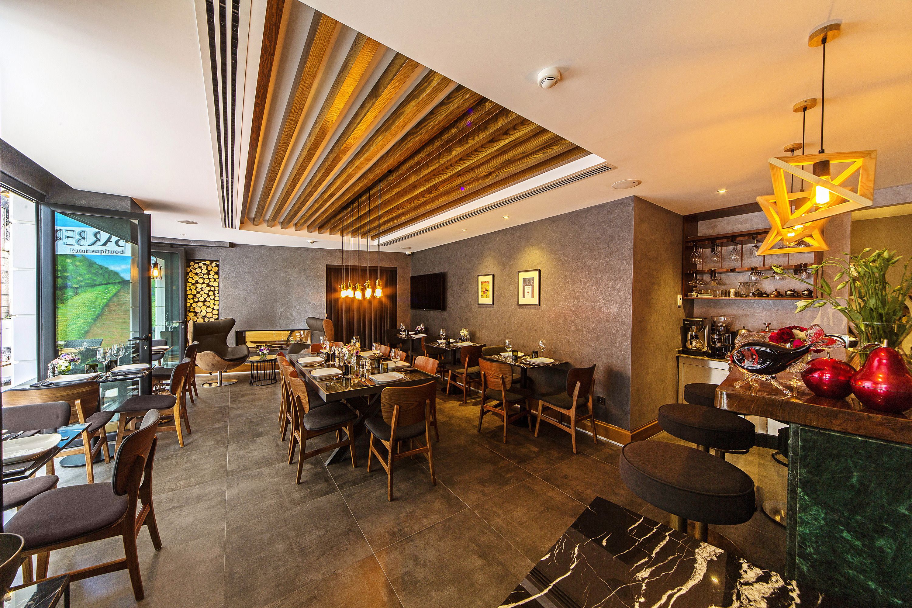 A modern restaurant with wooden ceiling panels, a bar area with stools, and a view of a green landscape through large windows.
