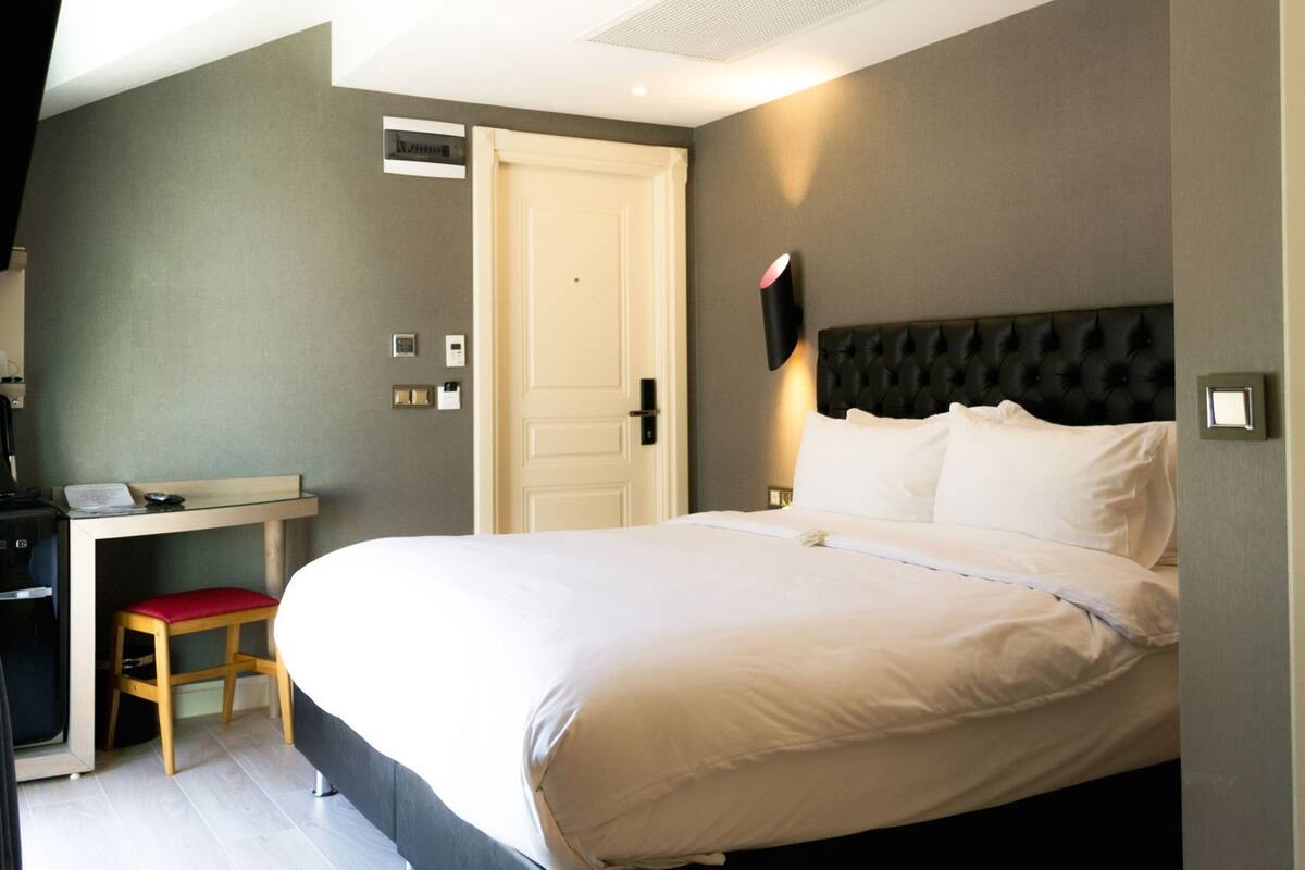 A hotel room with a bed, a desk, a chair, a door, and a wall-mounted light fixture.