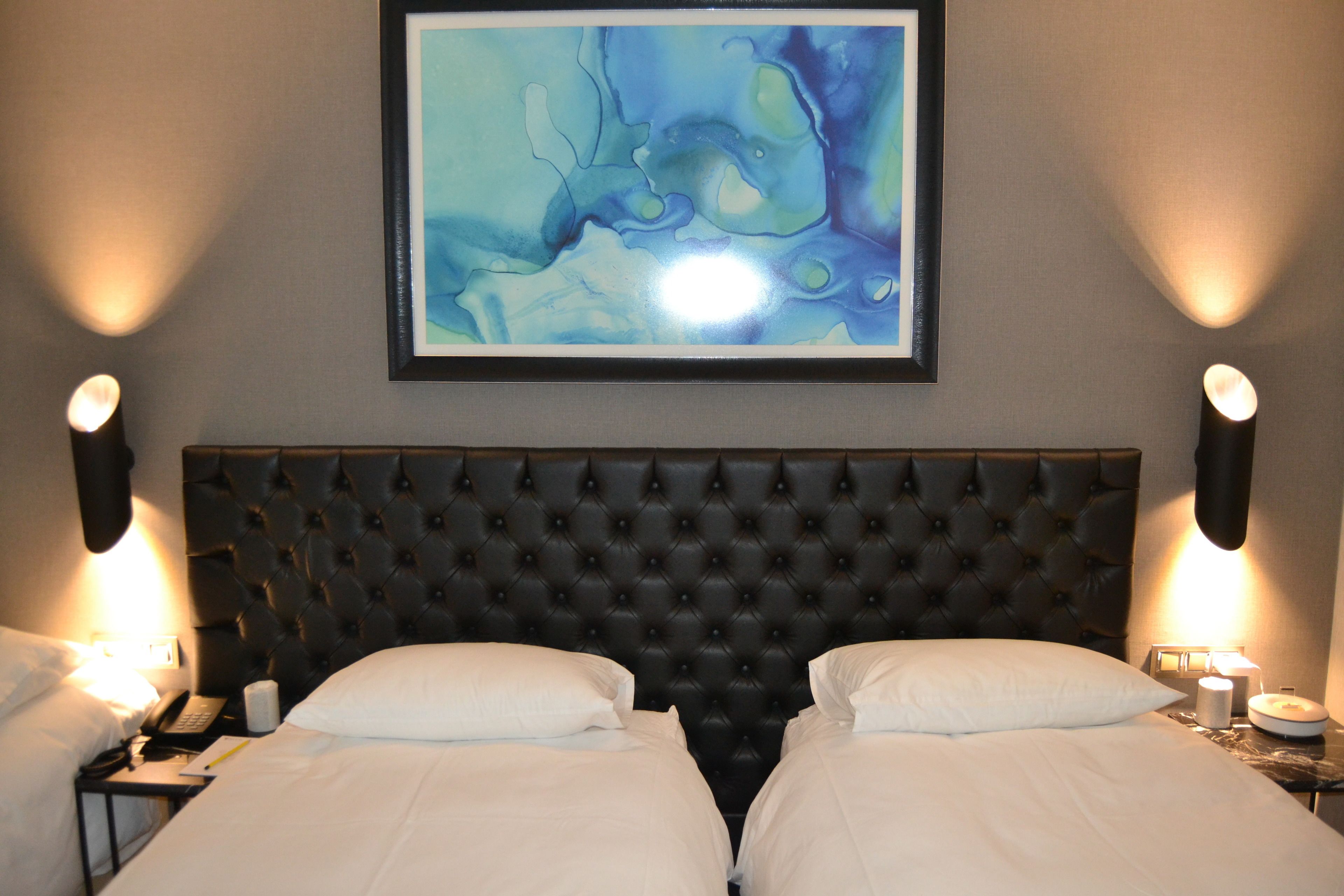 A hotel room with a tufted headboard, two beds with white linens, a framed abstract painting above the headboard, and wall-mounted lamps on either side of the bed.
