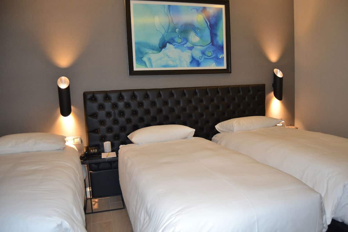 A hotel room with two single beds, a tufted headboard, bedside lamps, a framed abstract painting, and a nightstand with a lamp.