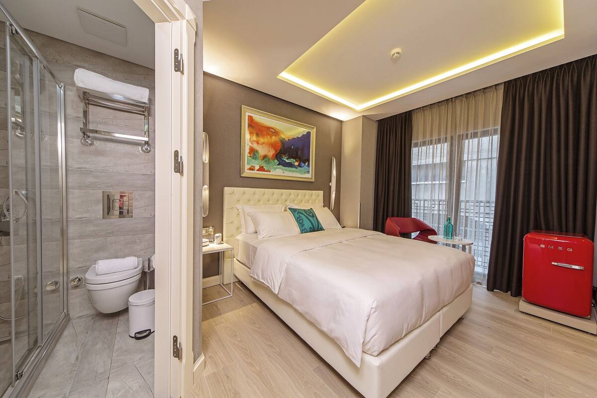 A modern hotel room with a large bed, a shower, a toilet, and a red refrigerator.