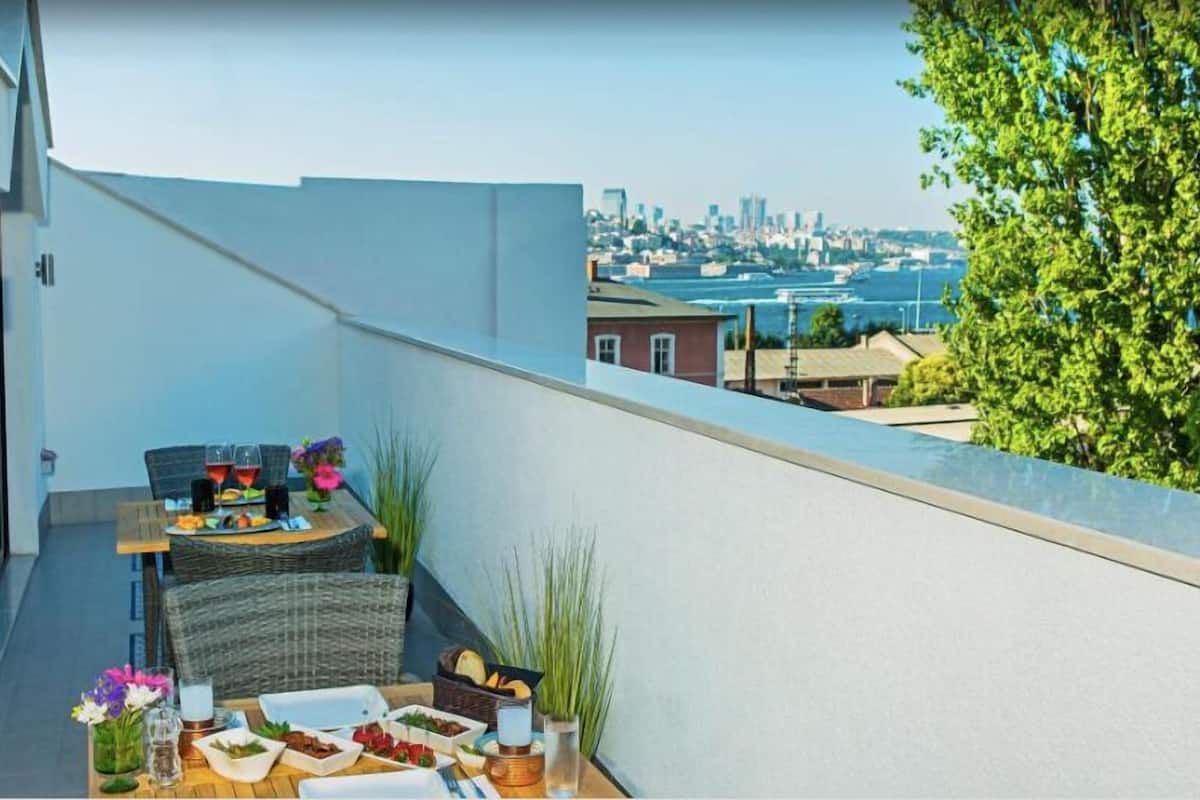 A balcony with a table set for dining, a wicker chair, and a cityscape view in the background.