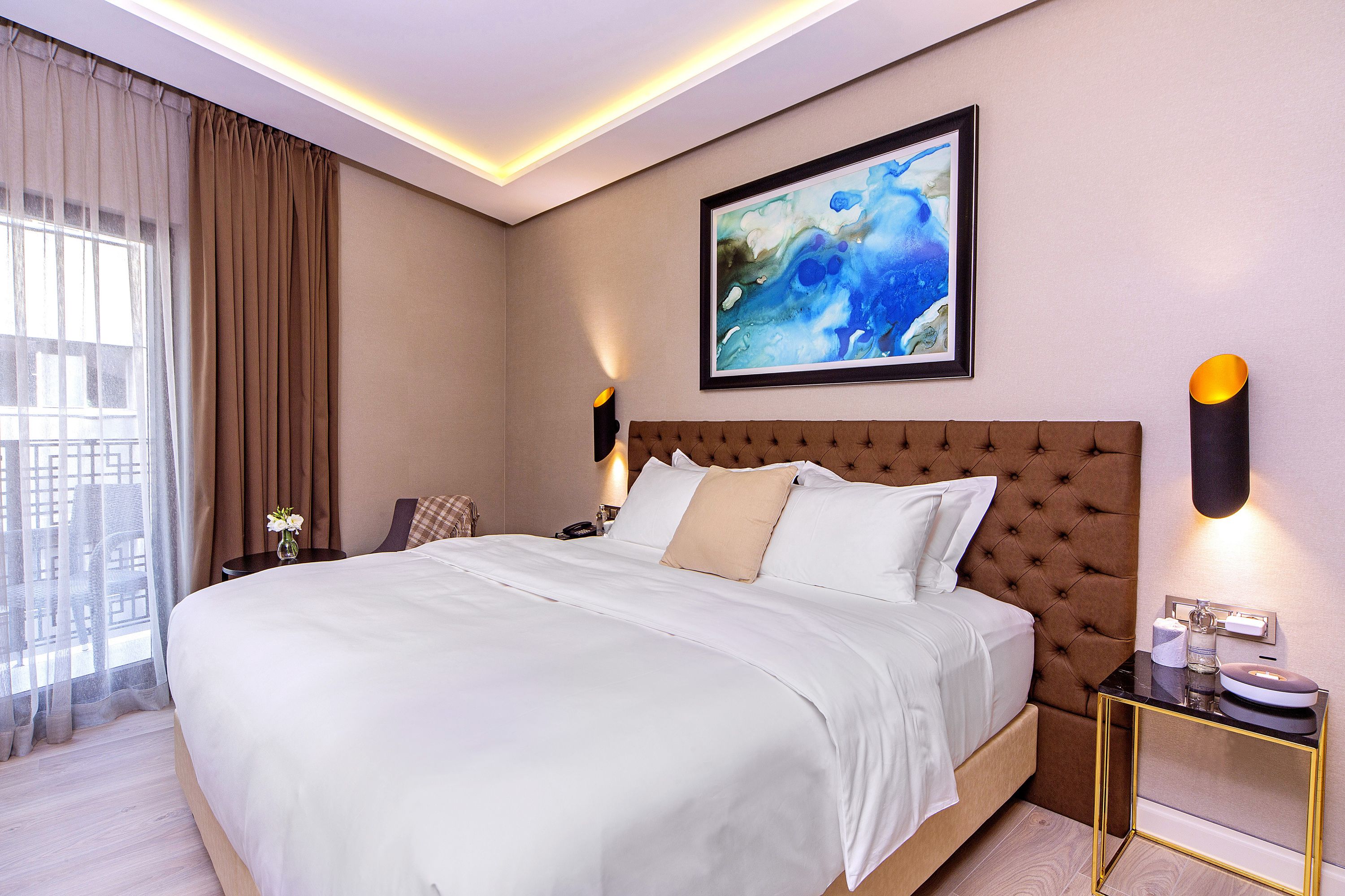 Quarto de hotel moderno com uma cama grande, uma mesa de cabeceira, um criado-mudo, um quadro abstrato emoldurado e uma janela com cortinas.