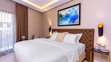 Quarto de hotel moderno com uma cama grande, uma mesa de cabeceira, um criado-mudo, um quadro abstrato emoldurado e uma janela com cortinas.