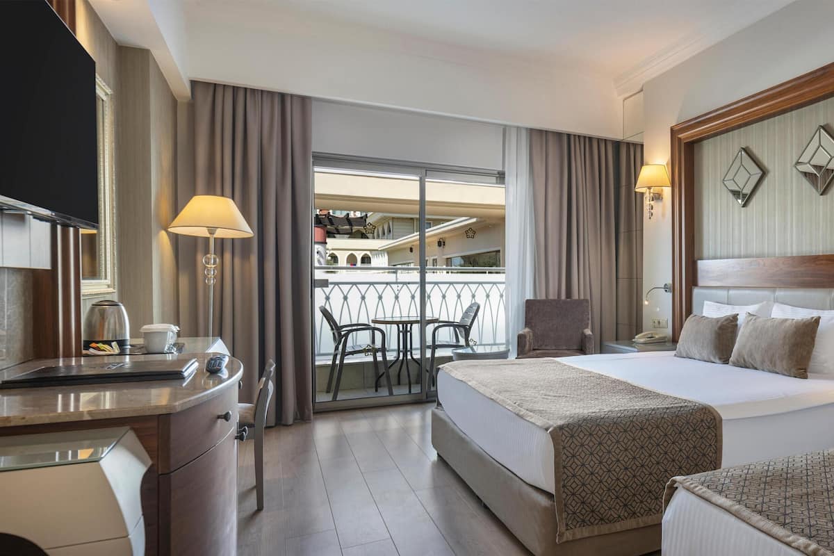 Habitación de hotel con dos camas, un escritorio, un balcón con sillas y un televisor.