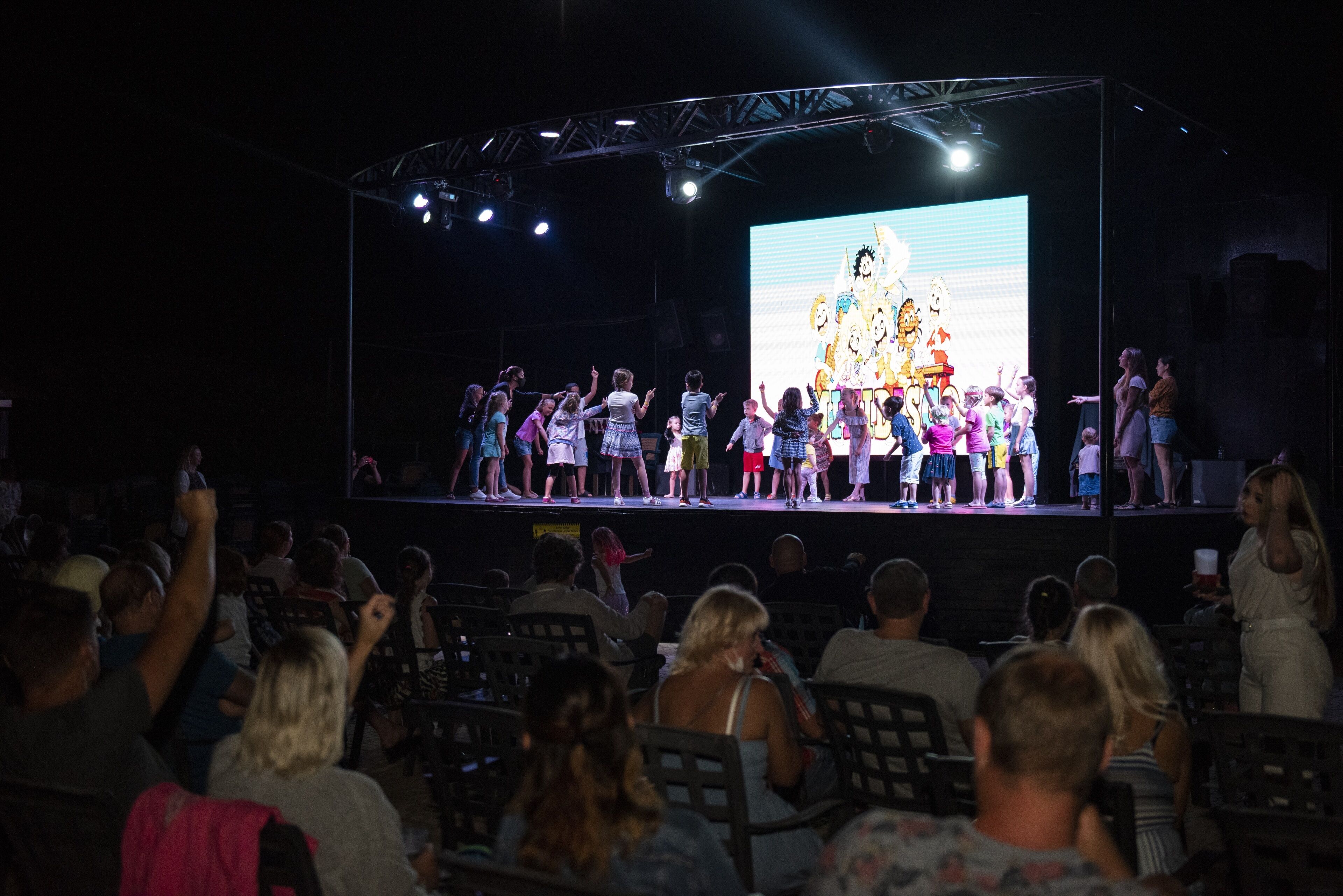 A stage with a group of people standing on it, a large screen displaying colorful characters, and an audience seated in front.