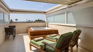 A sunlit balcony with a wooden bathtub, two green cushioned chairs, and a small table.