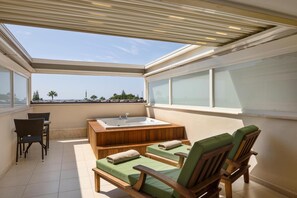 A sunlit balcony with a wooden bathtub, two green cushioned chairs, and a small table.
