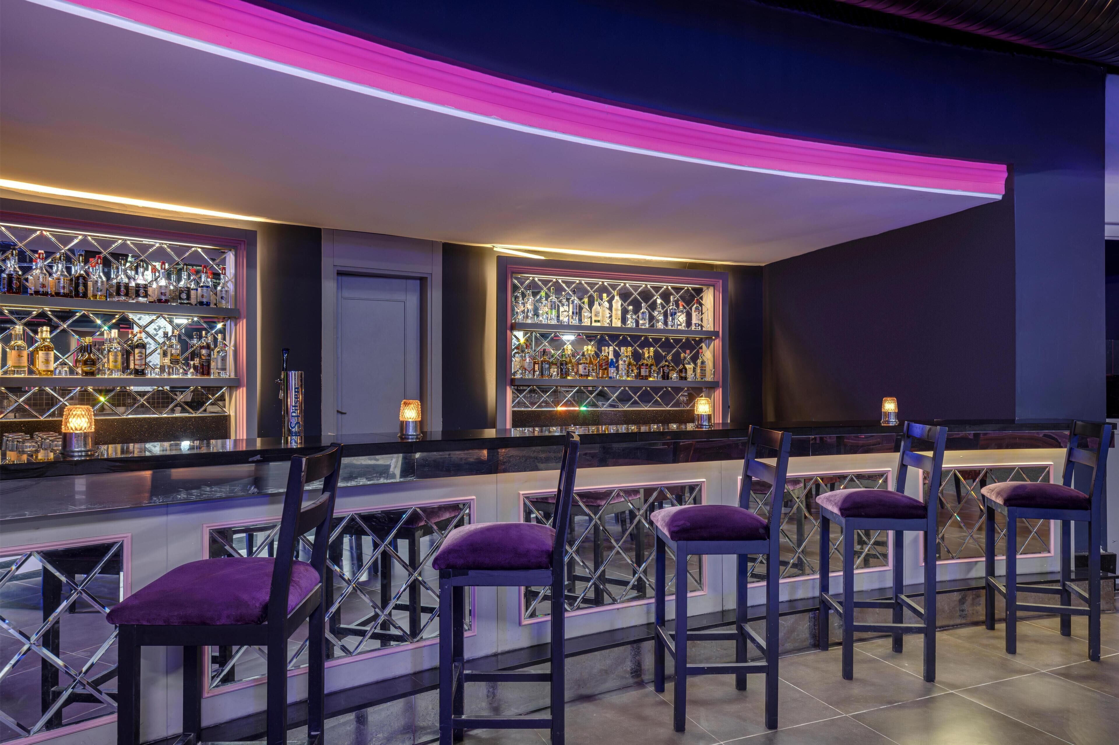 Uma área de bar com várias prateleiras de garrafas, banquetas e uma faixa de luz neon.