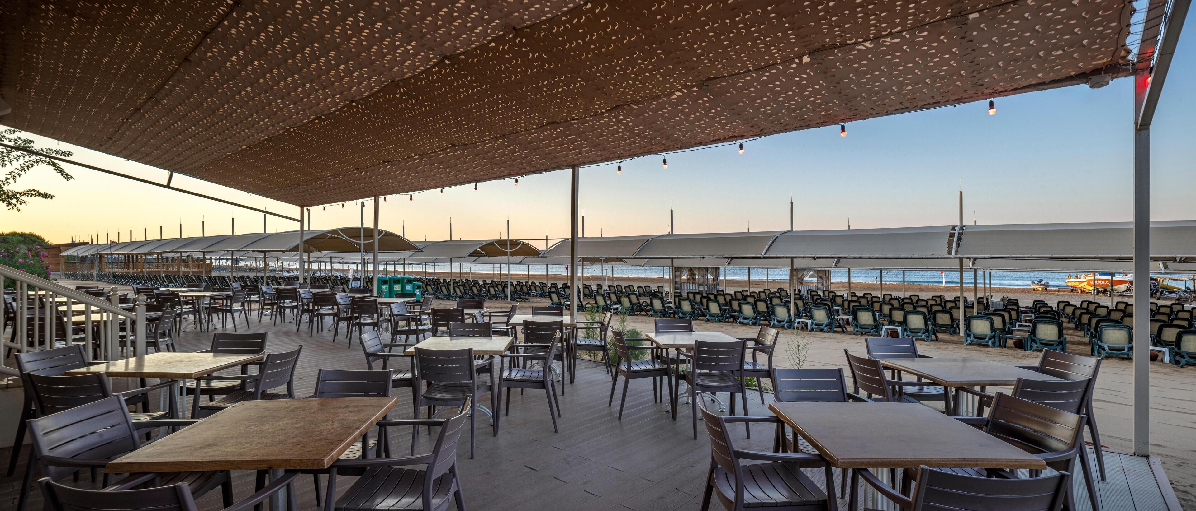 An outdoor seating area with tables and chairs, a covered section with a patterned roof, and a view of the beach in the background.