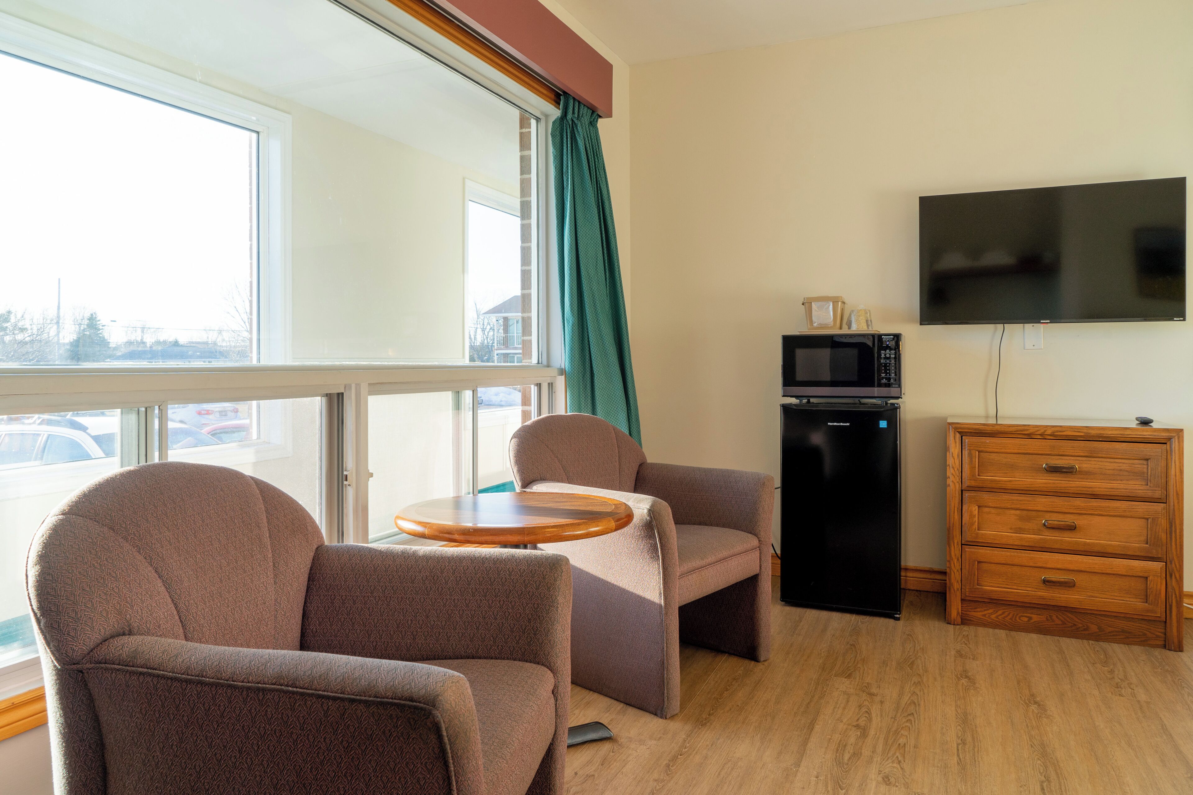 A room with a large window, a flat-screen TV, a microwave, a wooden dresser, and two armchairs.