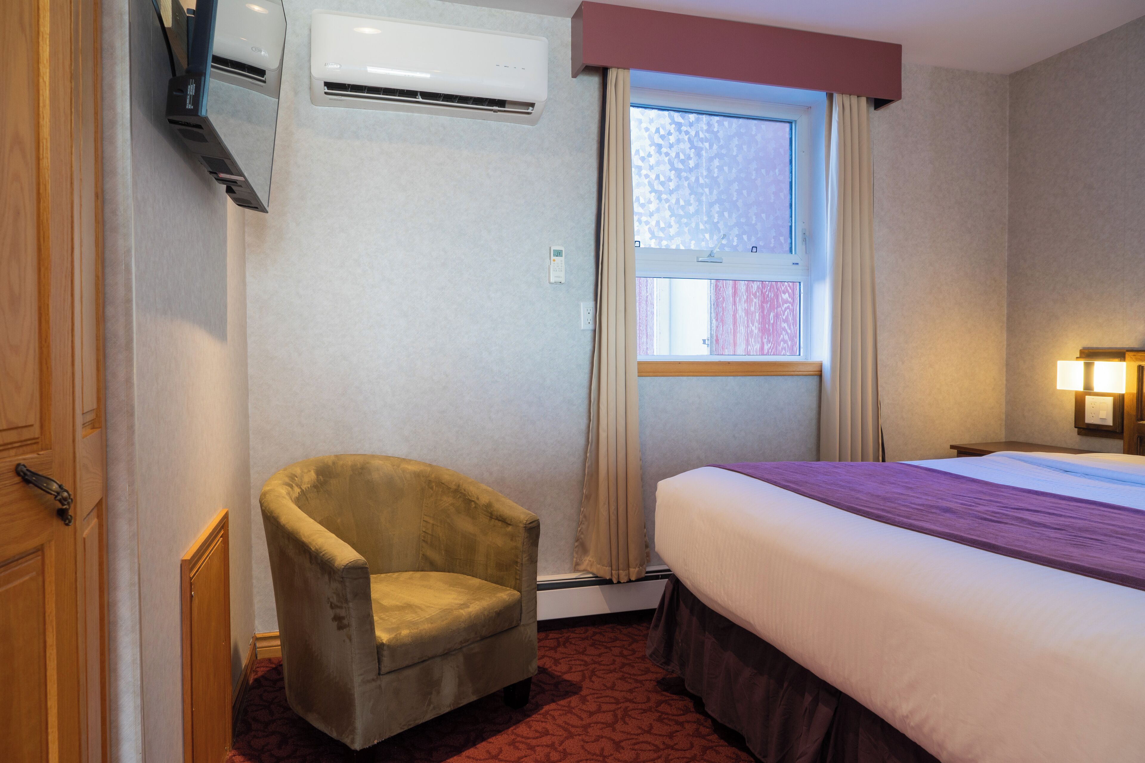 A hotel room with a bed, a chair, an air conditioning unit, a window with curtains, and a wall-mounted lamp.