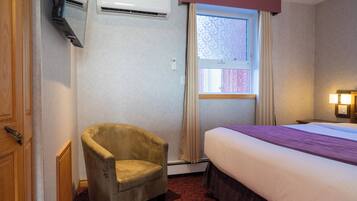 A hotel room with a bed, a chair, an air conditioning unit, a window with curtains, and a wall-mounted lamp.