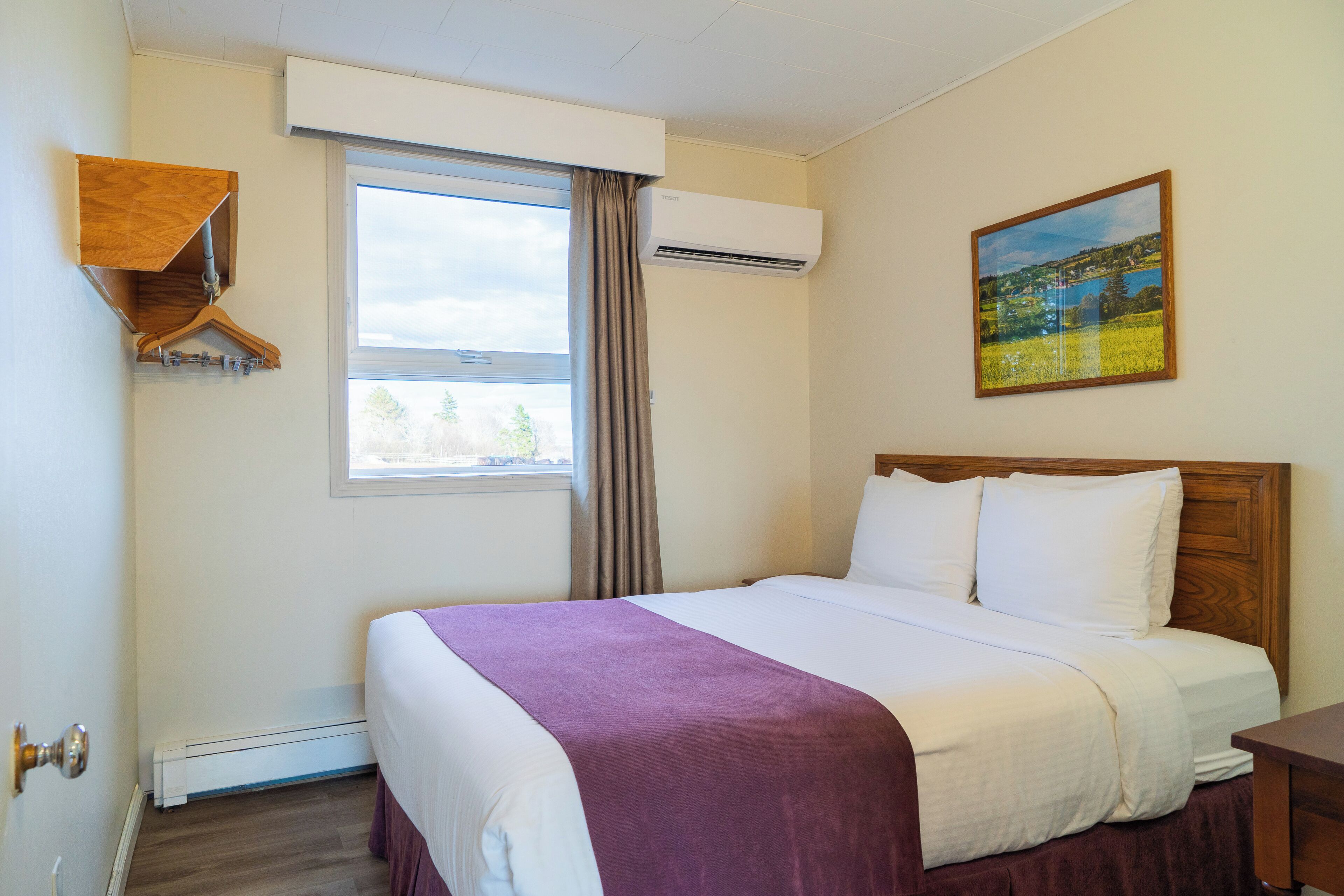 A hotel room with a bed, a window, an air conditioner, a painting, and a wooden shelf.