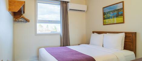 A hotel room with a bed, a window, an air conditioner, a painting, and a wooden shelf.