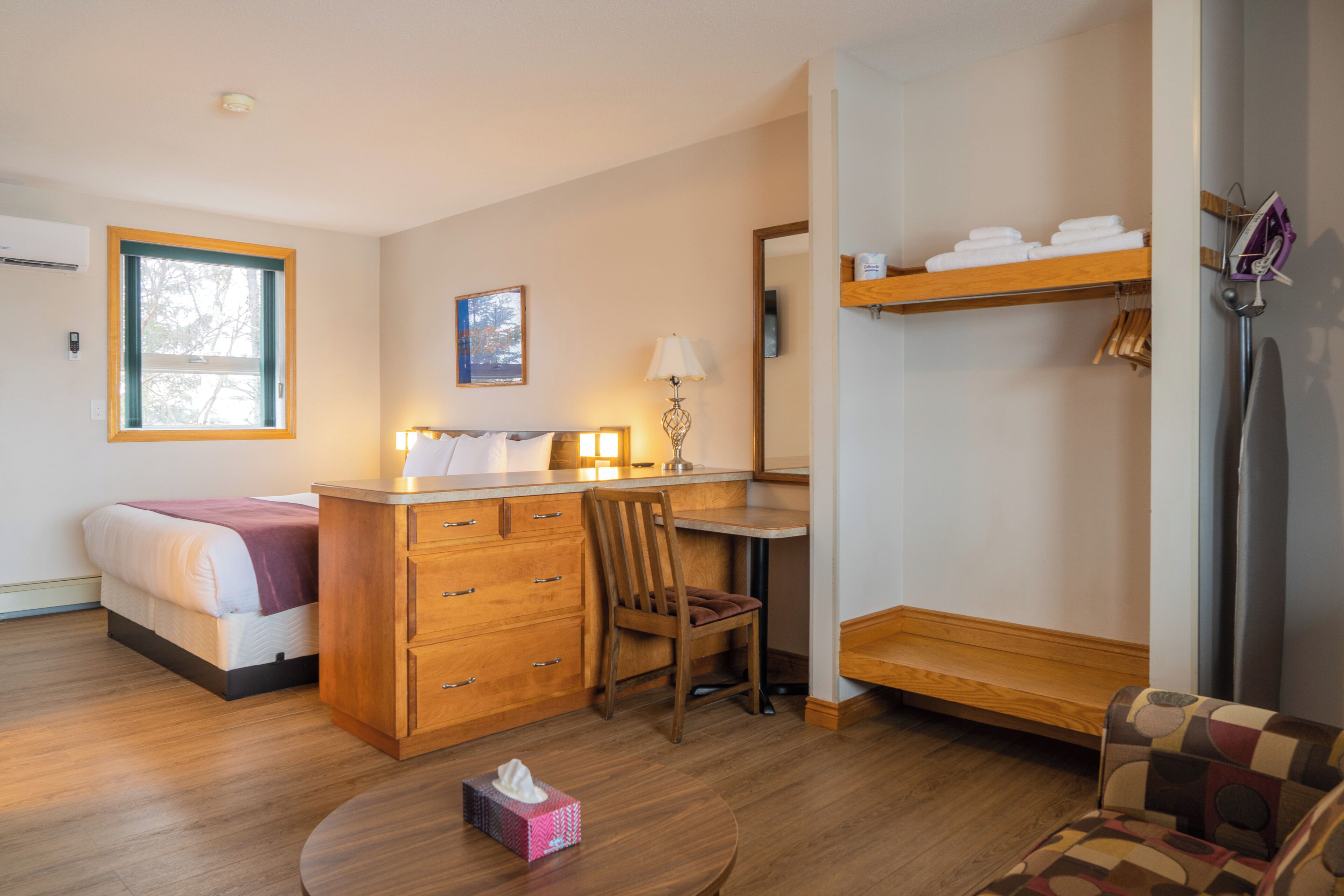 A hotel room with a bed, a wooden desk, a chair, a small table with a tissue box, a mirror, and a shelf with towels.