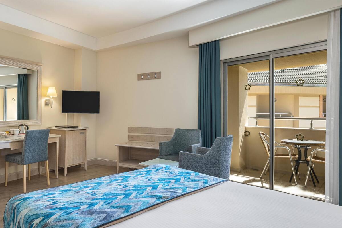 A hotel room with a bed, desk, chairs, a television, and a balcony with a table and chairs.
