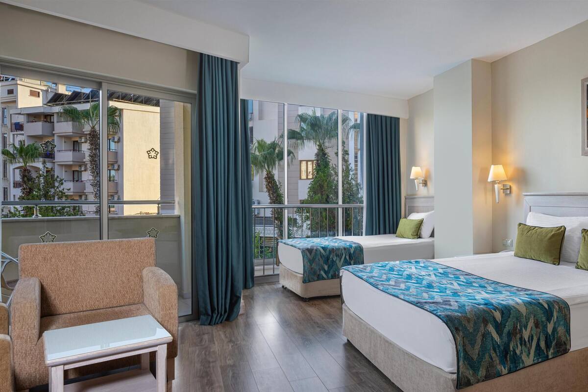 A hotel room with a large bed, two bedside tables, a chair, a window with curtains, and a view of the outside balcony.