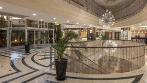 Un vaste atrium doté d’une rambarde centrale en verre, d’un grand lustre et de plantes en pot.