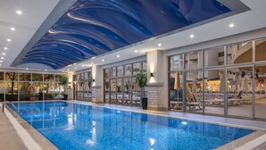 An indoor swimming pool with clear blue water, surrounded by glass walls and a ceiling with a blue wave design. - Side Star Park - All Inclusive (Manavgat)