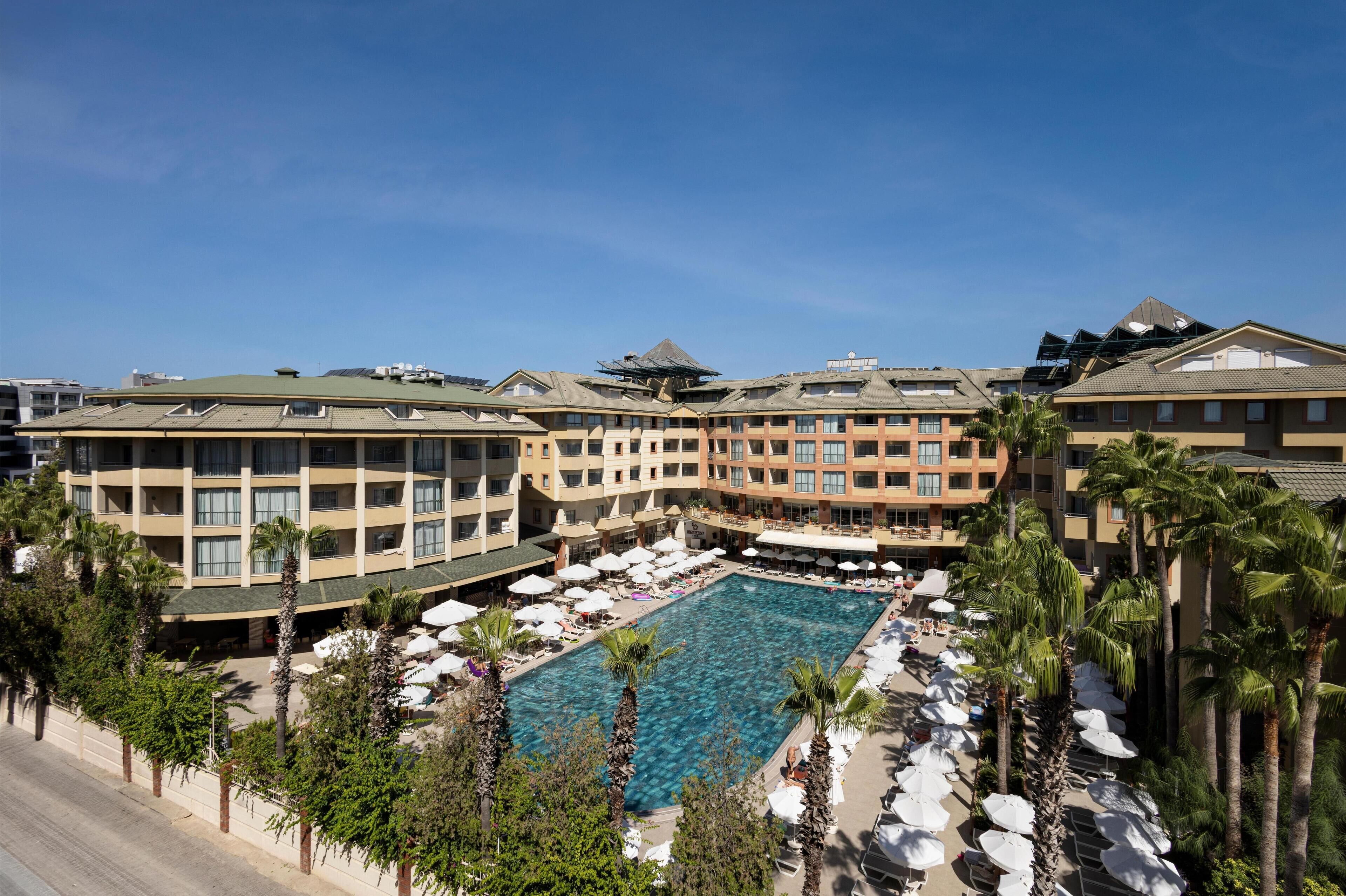 A hotel with a central swimming pool surrounded by palm trees and sun loungers.
