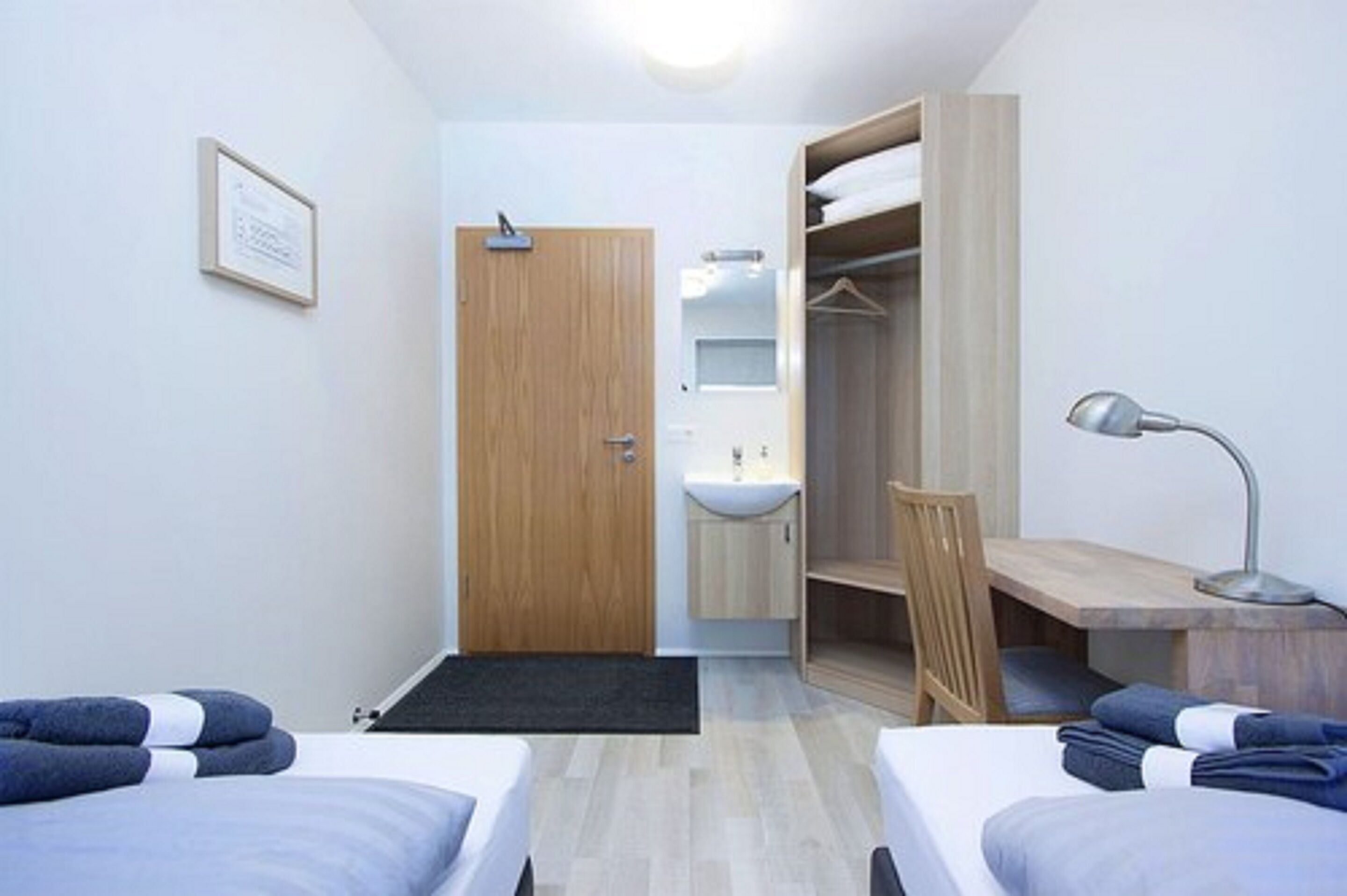 A small, modern hotel room with two single beds, a wooden wardrobe, a desk with a chair, a desk lamp, and a small bathroom with a sink and mirror.
