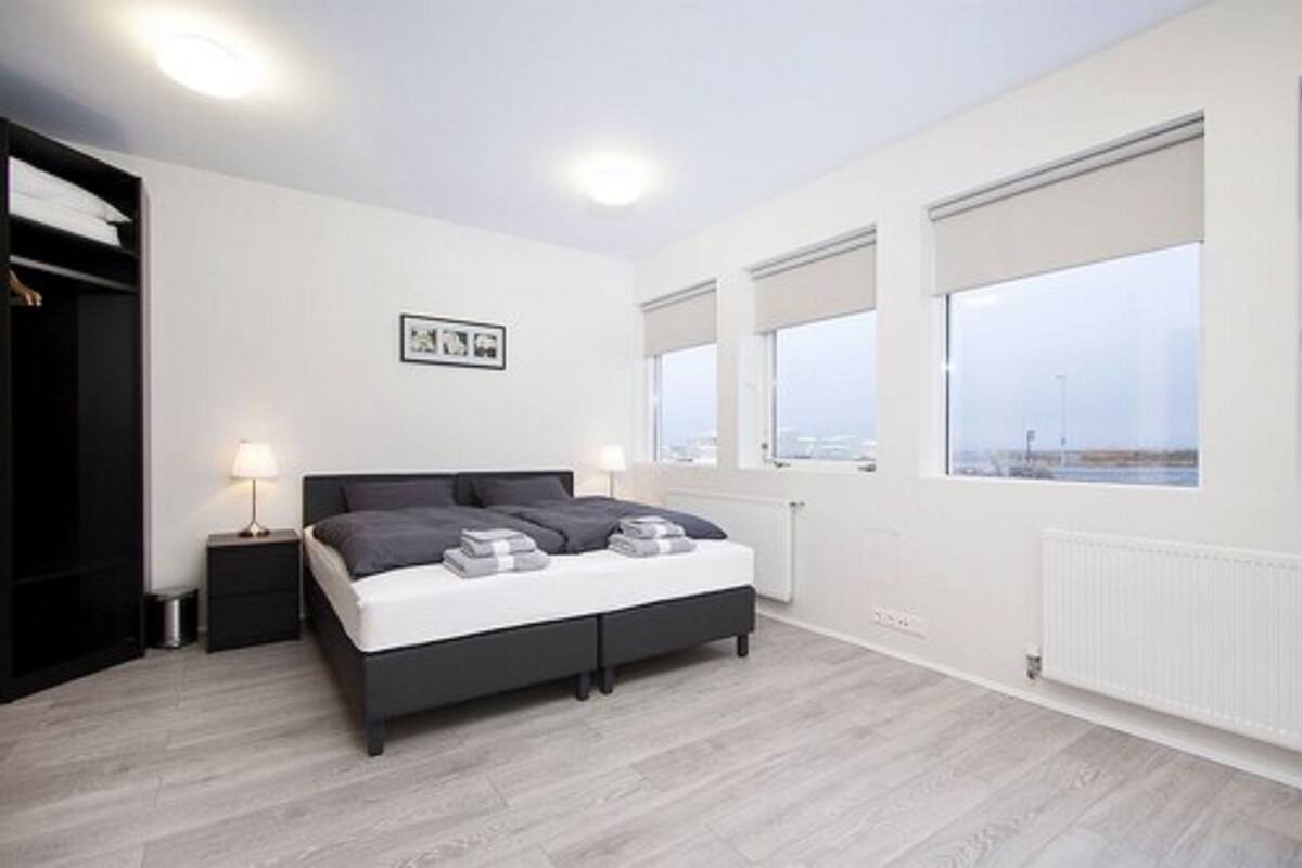 A modern bedroom with a bed, bedside tables, and a view through large windows.