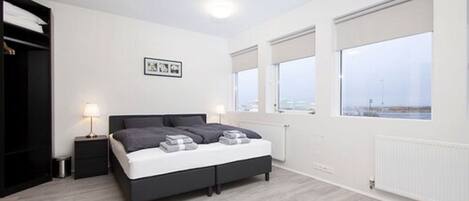 A modern bedroom with a bed, bedside tables, and a view through large windows.