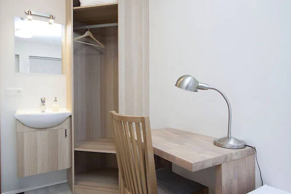 A small, modern bathroom with a wooden vanity, a mirror, a hanging shower curtain, and a desk with a lamp.