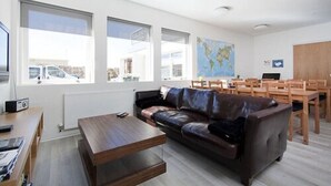 A modern living room with a leather sofa, wooden coffee table, and dining area with wooden chairs and a table. Large windows provide natural light and views of the outside.