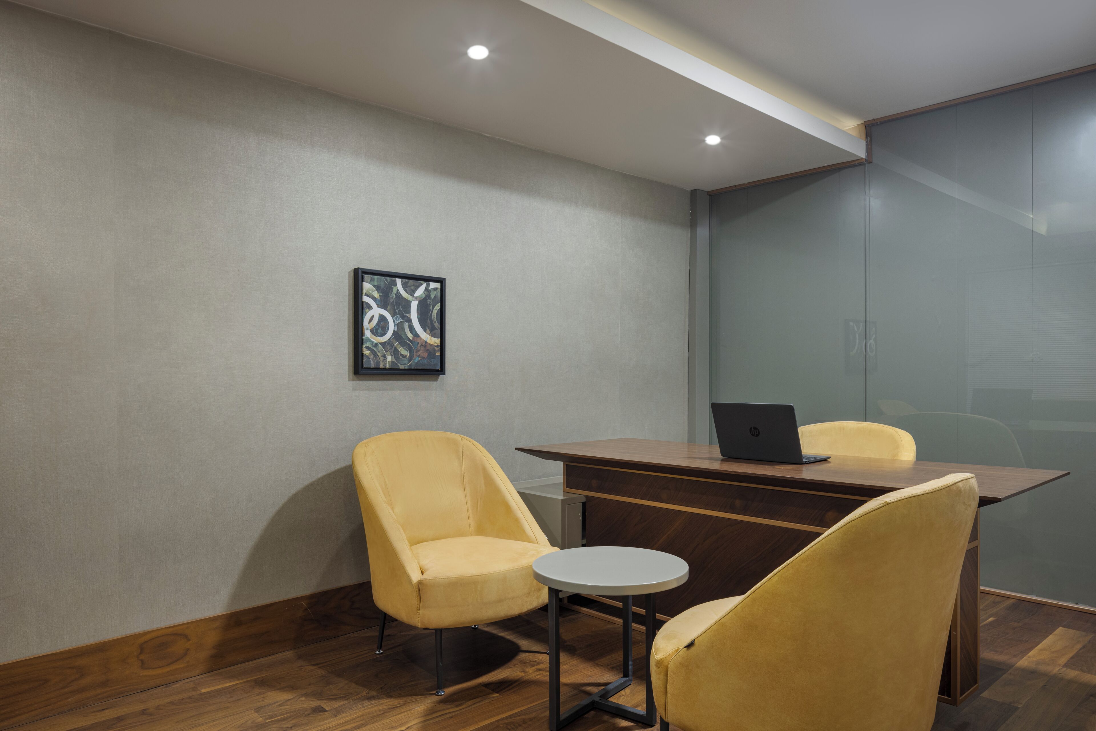 A conference room with a wooden desk, two yellow chairs, a laptop, and a framed abstract artwork on the wall.