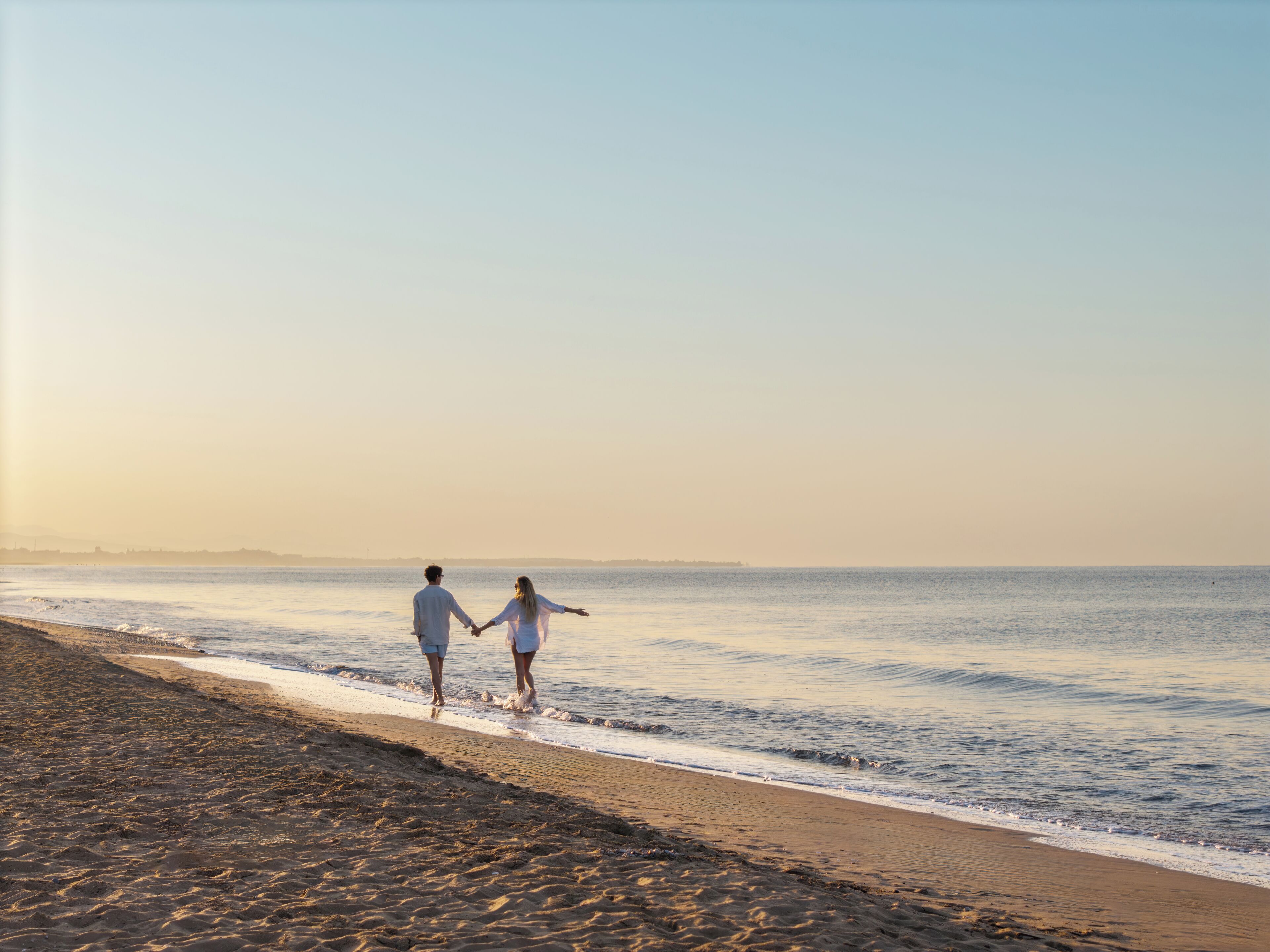 A couple walking hand in hand along a sandy beach with the ocean in the background.