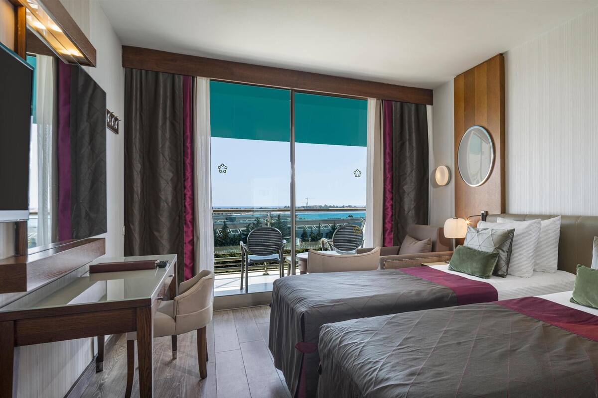 A hotel room with two beds, a desk, a chair, a large window with a view, and a mirror.
