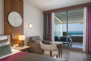 A hotel room with a bed, a desk, a chair, a round mirror, a lamp, and a view of the ocean through a sliding glass door.