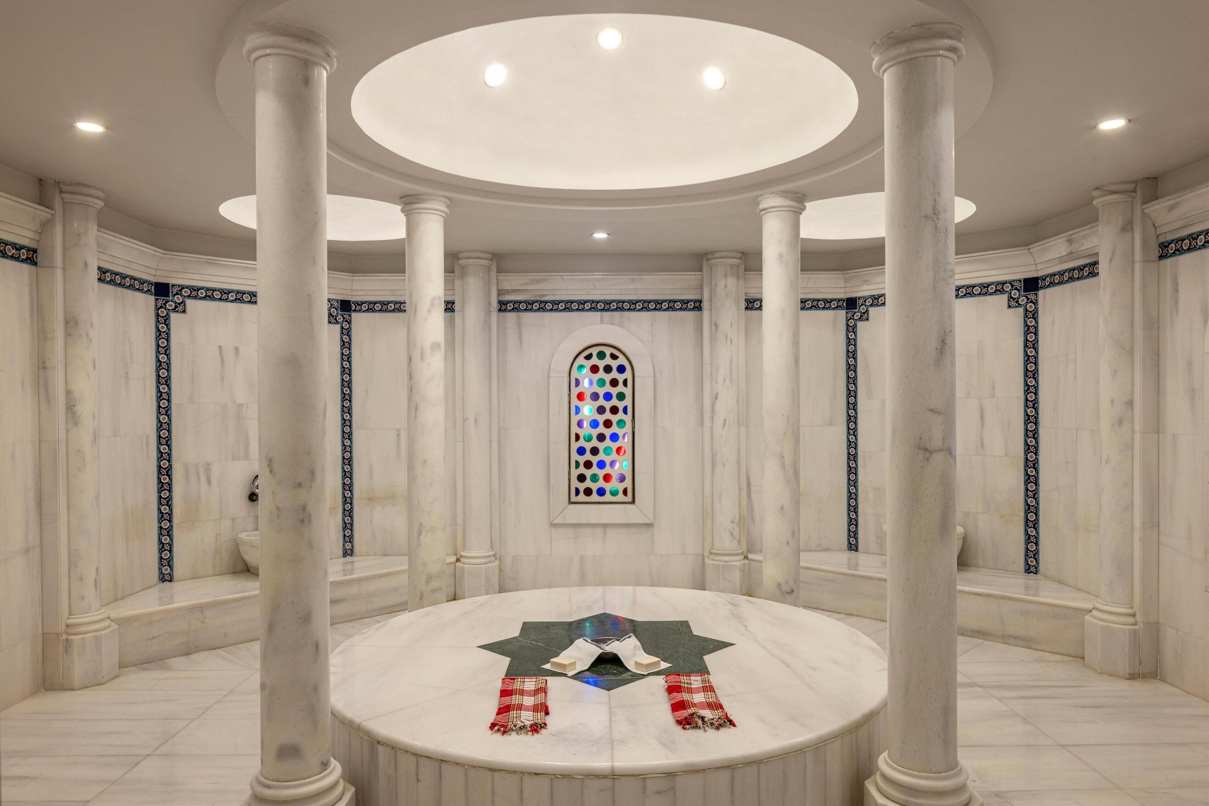 A circular marble room with a central star-shaped table, surrounded by tall columns, and a stained glass window.
