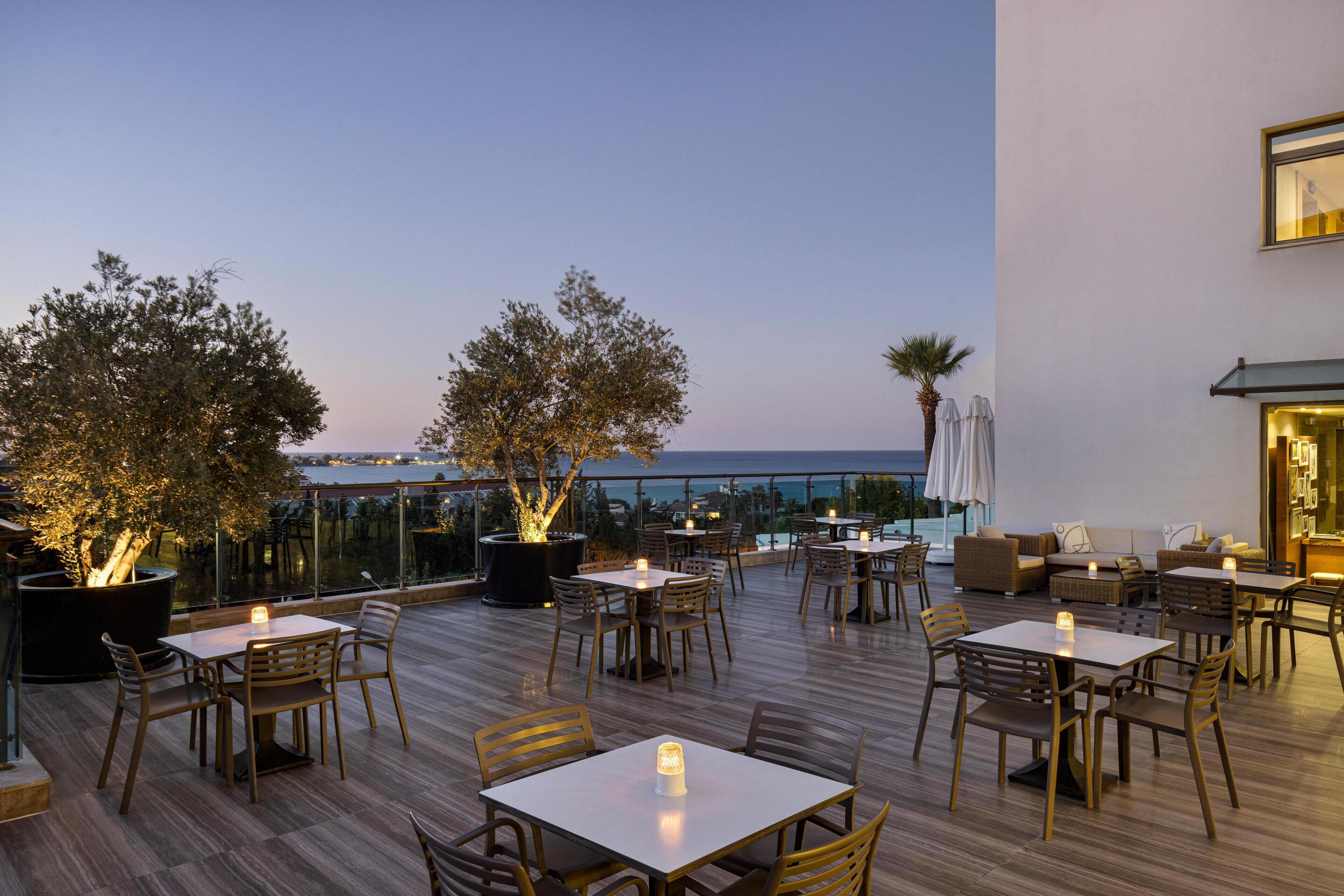 An outdoor dining area with tables and chairs, potted plants, and a view of the sea.