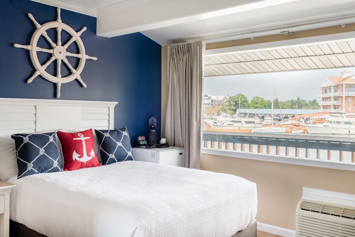 A bedroom with a bed, a ship's wheel decoration, a nightstand, and a view of boats through a window.