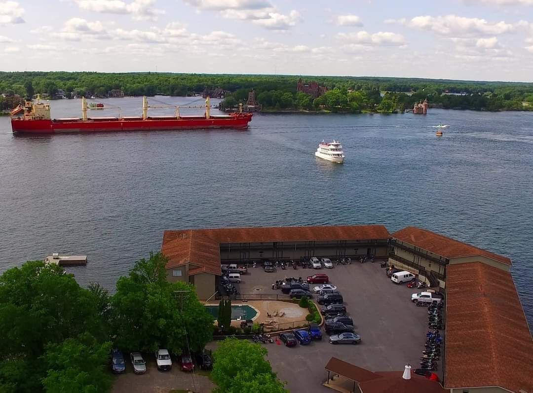 A large cargo ship and a smaller boat on a river. There is a parking lot with cars and a building with a red roof in the foreground.