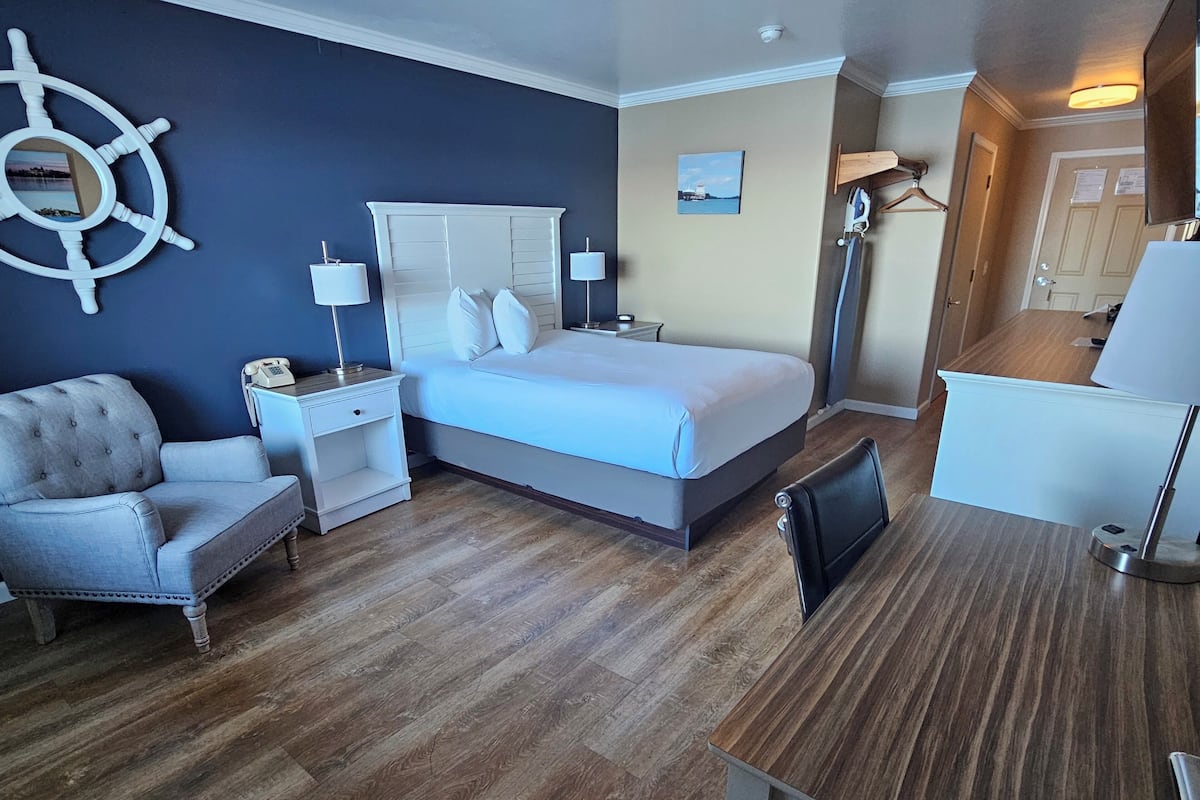 A hotel room with a bed, a chair, a desk, a lamp, a ship's wheel decoration, and a door.