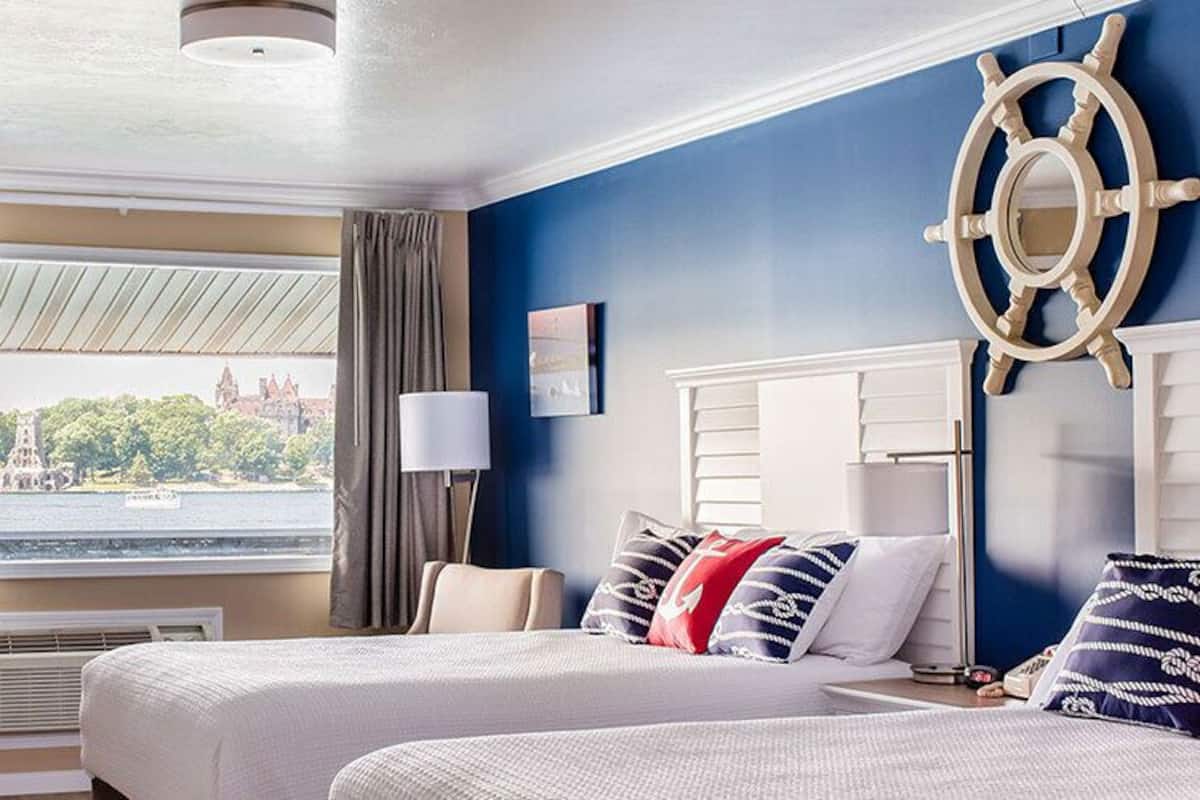 A hotel room with two beds, a large window with curtains, a ship's wheel decoration, and a television.