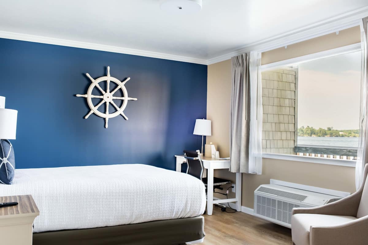 A hotel room with a large bed, a desk with a chair, a ship's wheel decoration, a window with a view, and an air conditioning unit.