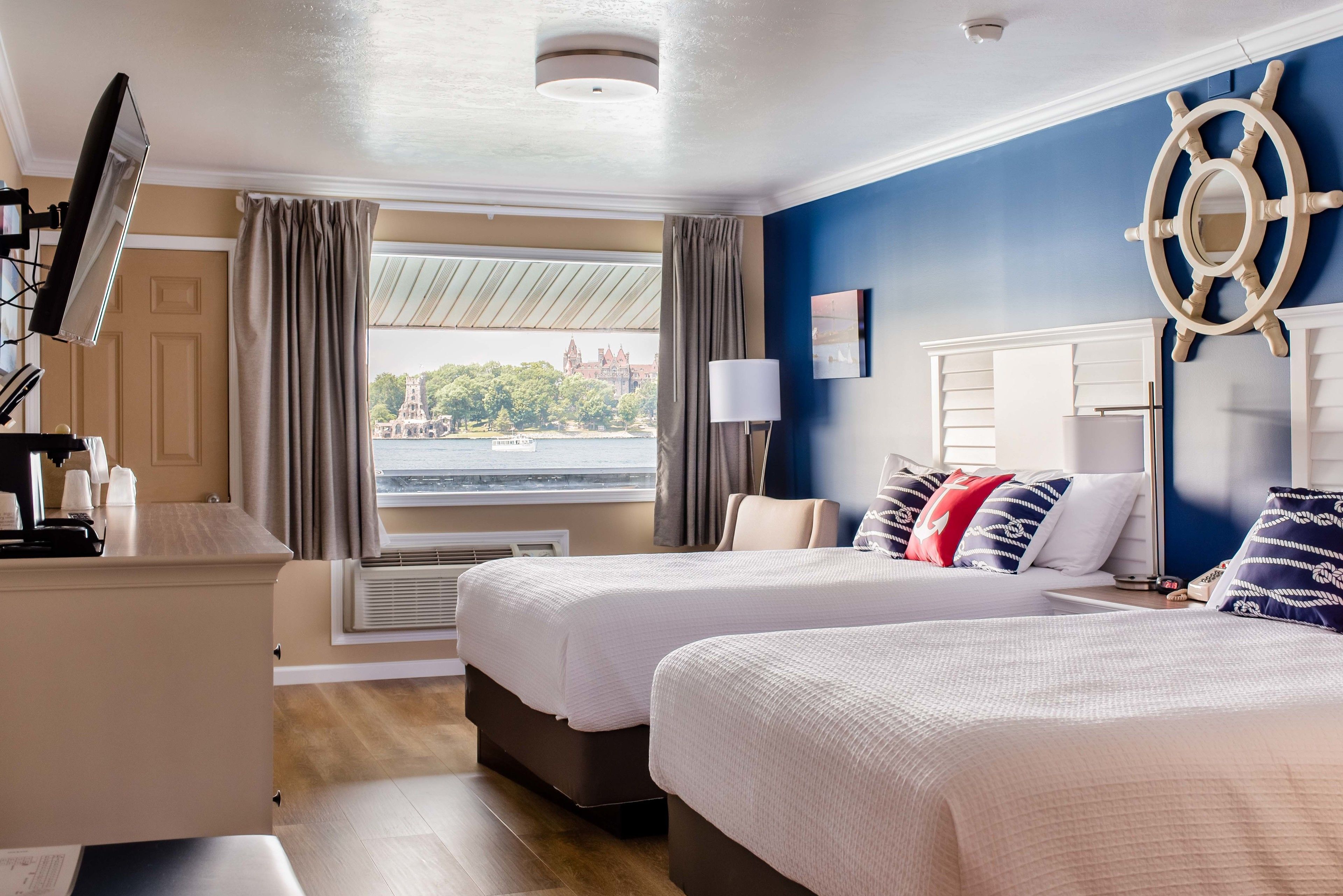 Ein Hotelzimmer mit zwei Betten, einem Schreibtisch, einem großen Fenster mit Vorhängen, einer Schiffslenken-Dekoration und Blick auf eine entfernte Skyline.