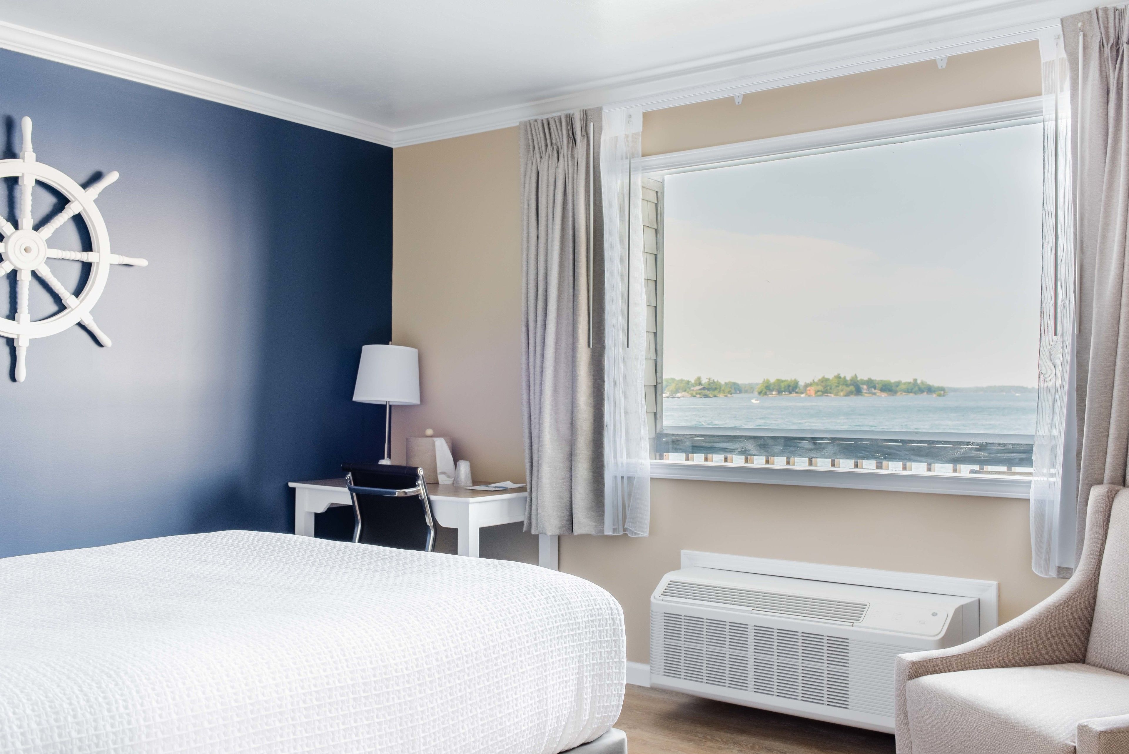 Een hotelkamer met een bed, een bureau, een stoel, een raam met uitzicht op de zee, en een schipperswiel decoratie.
