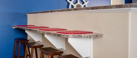 A bar area with a granite countertop, red beaded bar stools, and a white ship's wheel decoration on the wall.