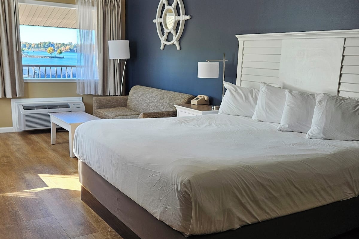 A large bed with white linens, a bedside table with a lamp, a ship's wheel decoration on the wall, and a view of a dock through a window.
