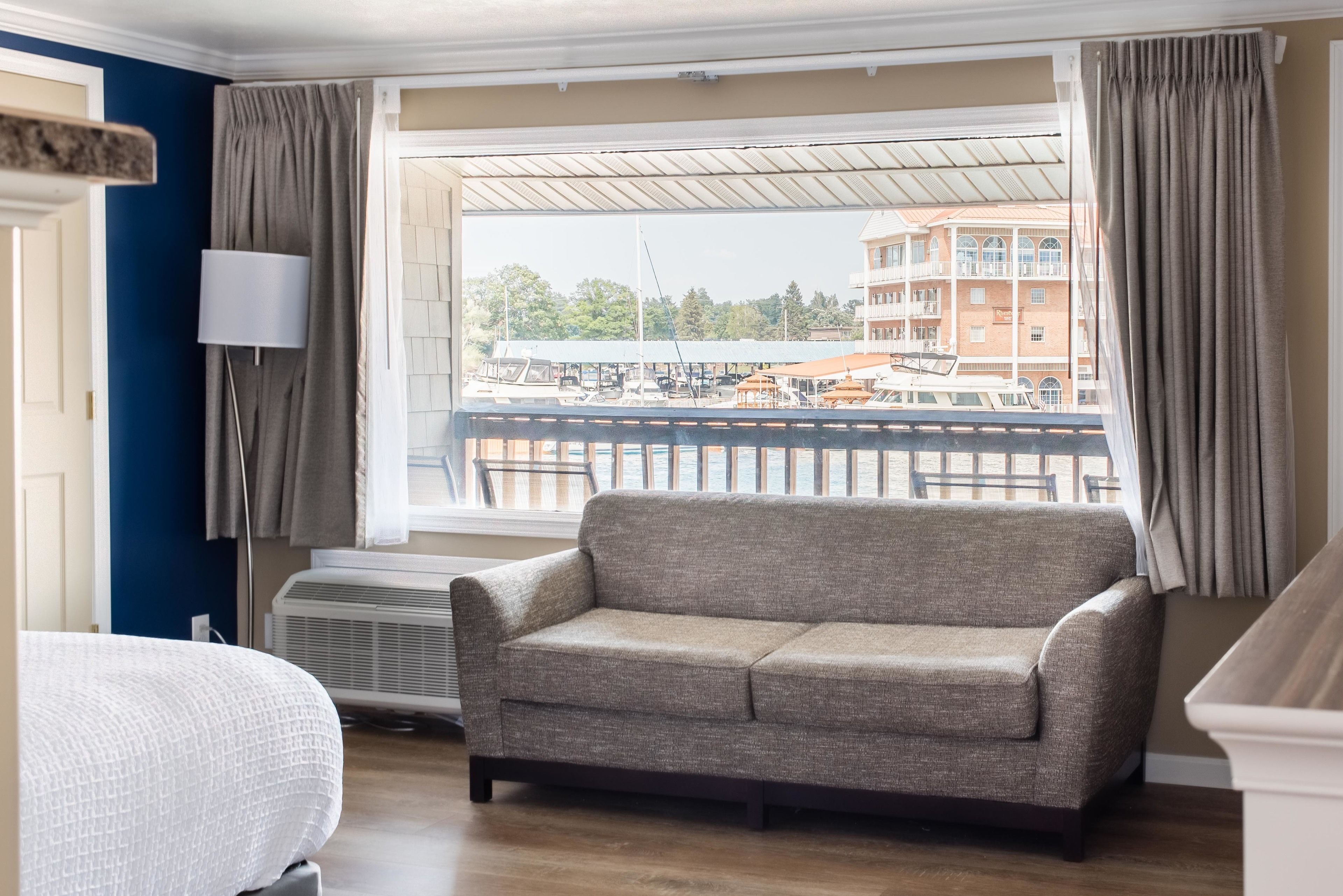 En stue med sofa, lampe og udsigt over en lysthavn med både og bygninger.