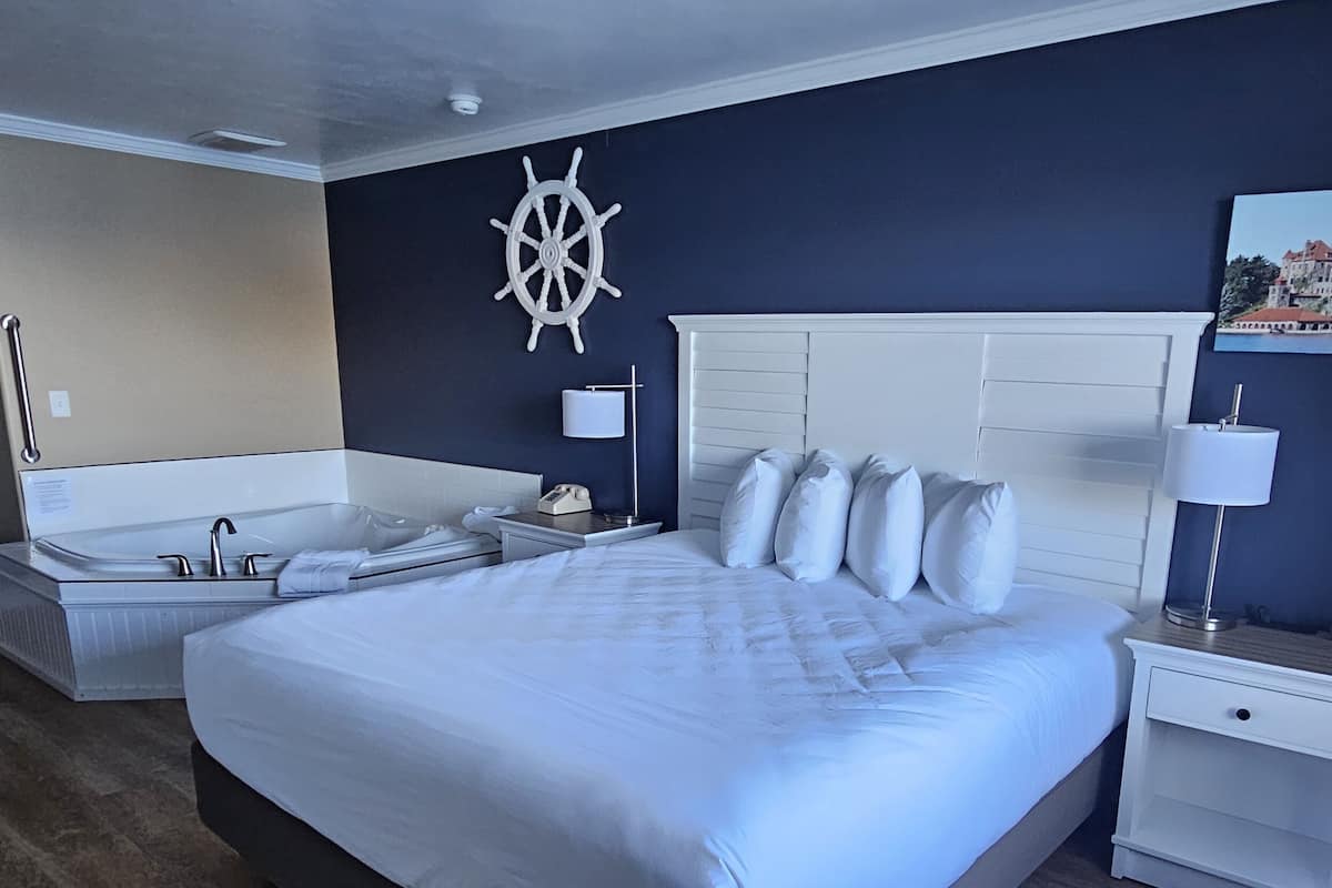 A hotel room with a large bed, a separate bathroom with a bathtub, a ship's wheel decoration on the wall, and a nightstand with a lamp.