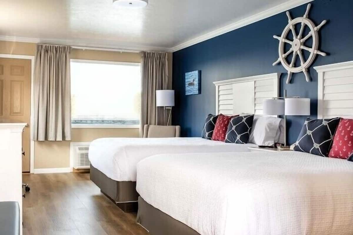 A hotel room with two beds, a large window with curtains, a ship's wheel decoration on the wall, and a view of the sea.